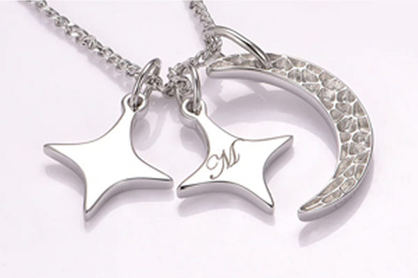 Silver crescent moon pendant with textured surface and two smooth star-shaped charms on a delicate chain, one star engraved with a cursive letter M, against a pale background.