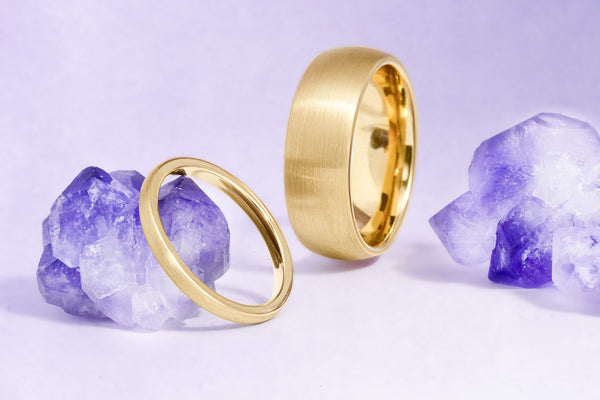 Minimalist gold couples ring set by Aquamarise, a slim women's band and wider brushed men's band beside raw amethyst crystals