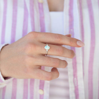 Oval white opal ring with two small round accent stones in a silver-tone setting, worn on a hand against a lilac striped shirt