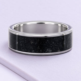 Black onyx wedding band with narrow silver tungsten edges, resting on matte lilac tiles under soft daylight.