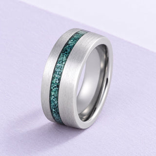 Brushed tungsten wedding band with an offset moss agate inlay, resting on fine lilac paper under soft daylight.