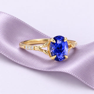 Yellow-gold oval blue sapphire leaf ring resting on a long dusty lavender silk ribbon, with clear accents and soft daylight.