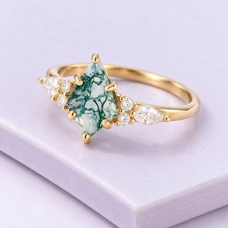 Yellow-gold Skye Kite moss agate engagement ring with clear accent stones resting diagonally on pale lilac porcelain tiles.