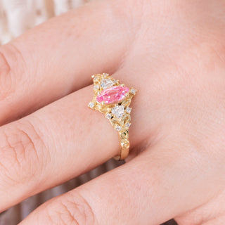 Yellow gold ring with marquise cut pink sapphire center stone and diamond accents on hand over a light background 