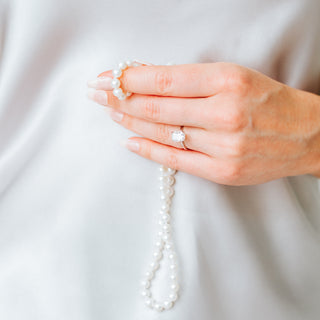 Hand holding a pearl necklace with a cushion cut diamond ring on a light background