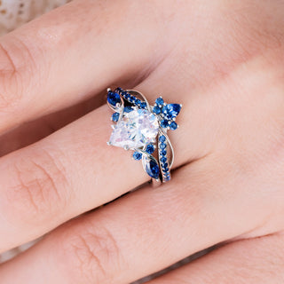 White gold engagement ring set with teardrop cut diamond center stone and lab-grown blue sapphire accents on hand over light background