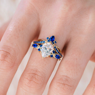 Yellow gold engagement ring set with teardrop cut diamond center stone and lab-grown blue sapphire accents on hand over light background