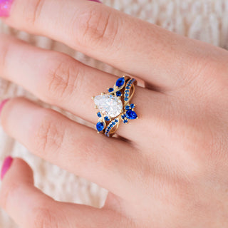 Yellow gold engagement ring set with teardrop cut diamond center stone and lab-grown blue sapphire accents on hand over light background