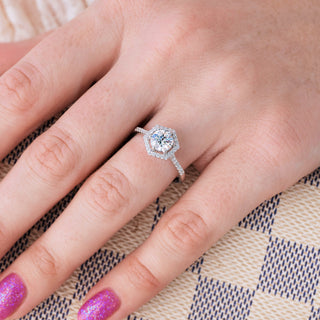 White gold engagement ring with round cut diamond center stone set in a hexagon setting on hand over a checkered background