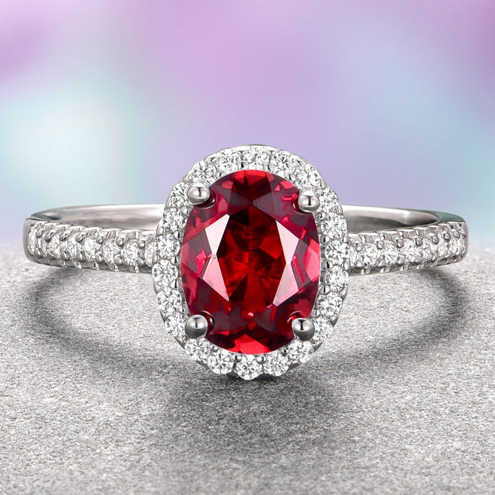An oval red garnet engagement ring with a halo of white gold and accent stones set on a textured metal band.