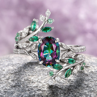 A sterling silver ring set featuring an oval Mystic Topaz center stone with leaf motifs adorned with emerald-green stones, displayed on a rock.
