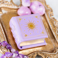 Dawn Court Leather Book Ring Box - Amethyst Light Edition (Small) in soft lavender with gold celestial design by Aquamarise, styled with amethyst stones on a decorative golden tray.