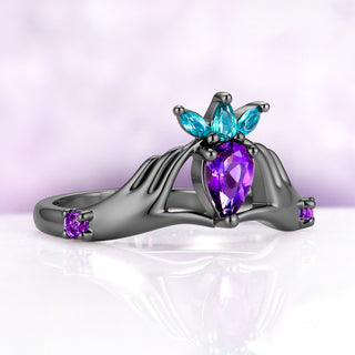 Three-quarter view of the amethyst Claddagh engagement ring in black ruthenium, showing the fingers of each hand wrapped around the stone