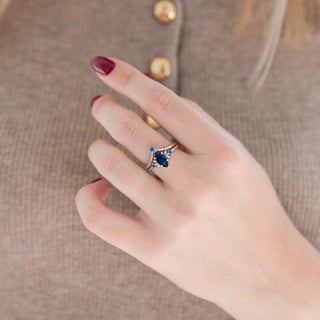 Silver marquise cut sparkly blue sandstone engagement ring with crescent moon motif and blue sapphire accent stones and chevron wedding band with blue sapphire held in hand against warm brown background