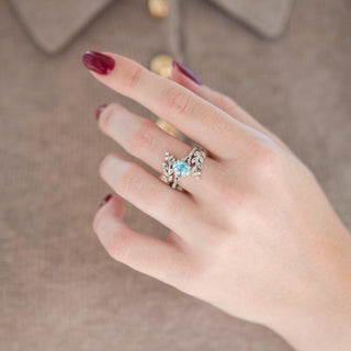Silver leaf guard wedding band with diamond and moonstone accents and oval aquamarine engagement ring displayed in hand against warm brown backdrop