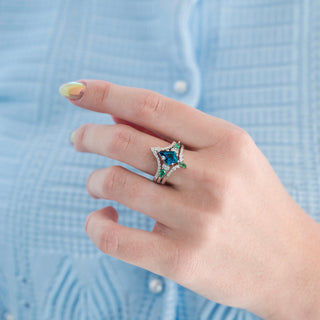 Hand wearing a star-shaped ring with blue and green gemstones against a light blue shirt background