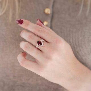 Rose gold pear/teardrop-cut dark red garnet engagement ring with diamond accent stones and curved wedding band with diamonds held in hand against warm brown background