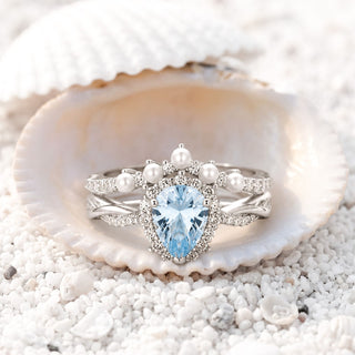Sterling silver aquamarine pear halo engagement ring with a pearl crown wedding band nestled in a seashell on white sand, Aquamarise collection banner