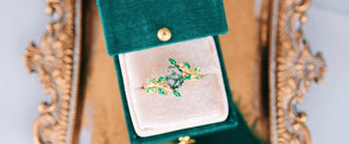 Gold and emerald ring in a green velvet box with an ornate gold frame.