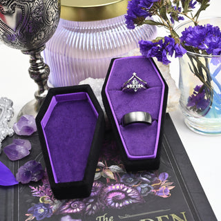 Black velvet coffin ring box with purple lining holding an engagement ring and a wedding band