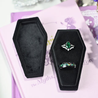 Black velvet coffin ring box holding an engagement ring and a wedding band, with the lid beside it