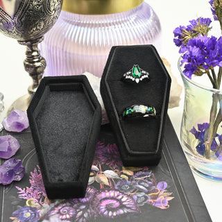 Open black coffin ring box showing the two ring slots and the velvet-lined lid, styled with amethyst and purple flowers