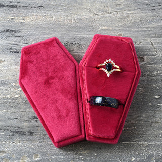Red velvet coffin ring box holding an engagement ring and a wedding band, with the lid beside it