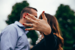 Woman showing an engagement ring on her left hand with red nail polish, while a blurred couple embraces in the background surrounded by greenery.