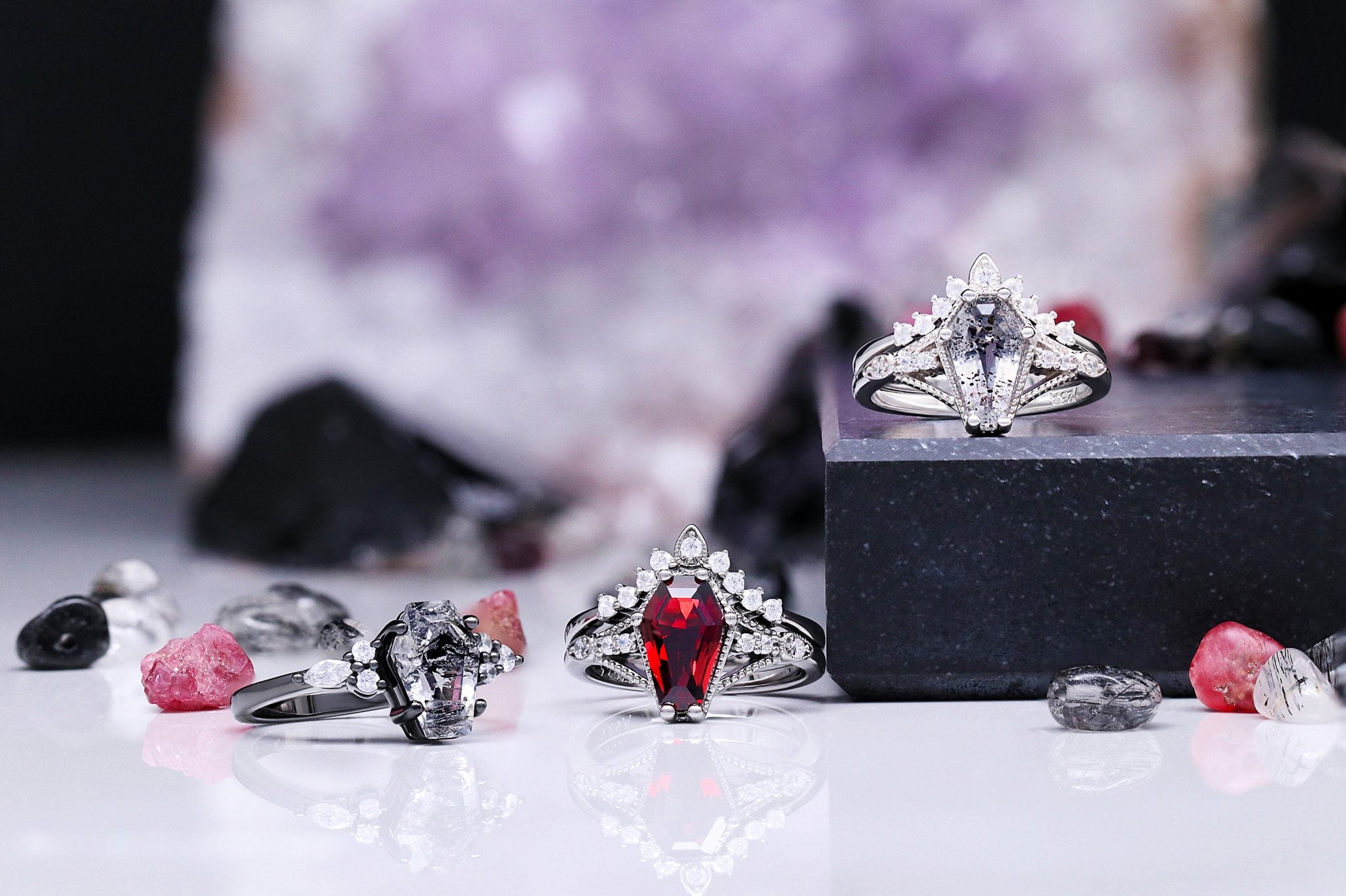 Pear-cut red garnet and rutilated quartz rings styled on black stone blocks with raw red crystals and an amethyst geode behind.