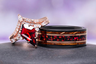 Rose gold ring with a vibrant red gemstone and small diamonds beside a black band featuring red stone inlays and wooden accents on a smooth dark surface against a soft purple backdrop.