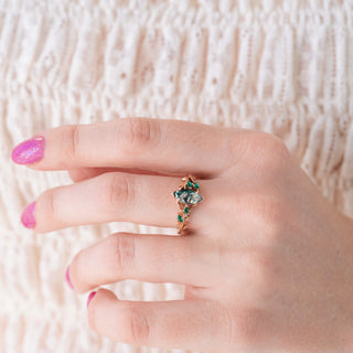 Close-up of a hand wearing a marquise moss agate and emerald ring on a neutral background