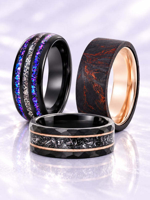 Three men's wedding bands on lilac satin: black band with galaxy opal and meteorite channels, black damascus-pattern band with rose gold interior and black hammered band with meteorite and rose gold stripes, Aquamarise banner