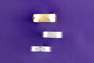 Wide view of three men's wedding bands on purple fabric: hammered gold, brushed silver, and a bark-textured band with a bezel-set diamond.