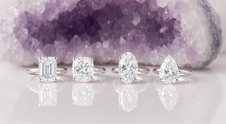 Four diamond rings on a reflective surface with a purple crystal formation in the background