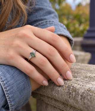 Hand in a denim jacket resting on a stone ledge wearing a kite-cut moss agate engagement ring with marquise accents, Aquamarise banner