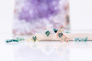 Three delicate rings with green moss agate stones in gold, silver, and rose gold settings, styled with matching green crystals and a soft purple background.