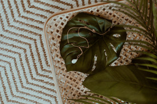 Delicate gold necklace with a teardrop pendant resting on a large green monstera leaf, placed on a woven surface beside tropical foliage and a textured rug.
