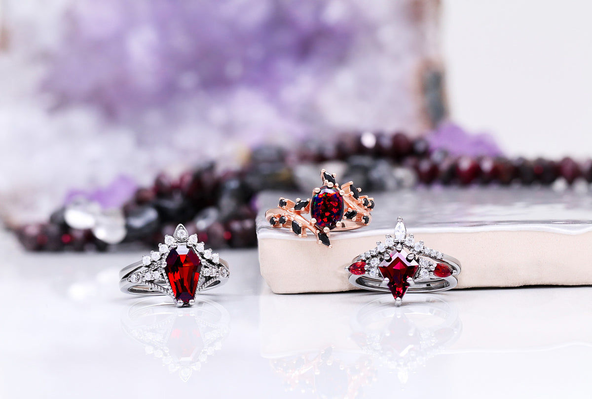 Three unique engagement rings featuring deep red gemstones with intricate silver and rose gold settings, displayed on a reflective surface with a soft purple crystal background.