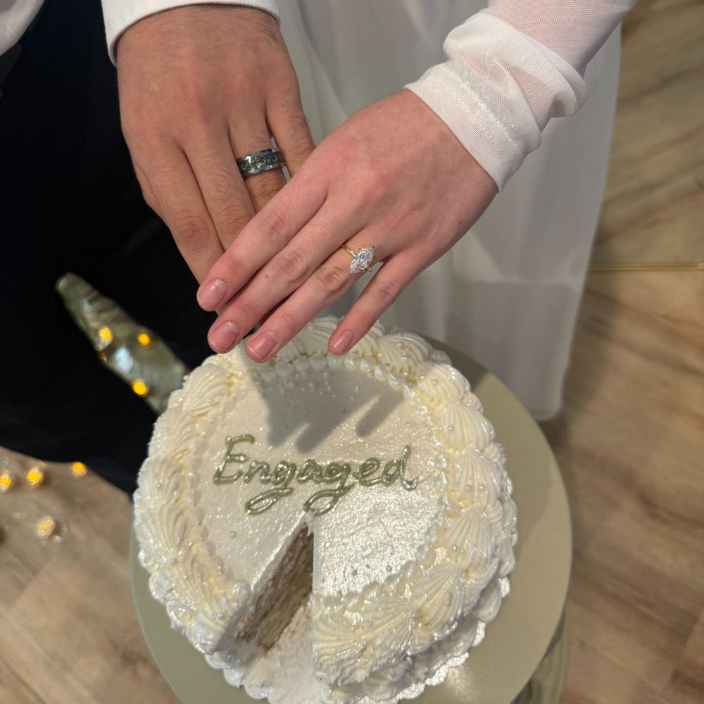Customer photo of a newly engaged couple resting their hands on an Engaged cake, showing an Aquamarise engagement ring and men's band