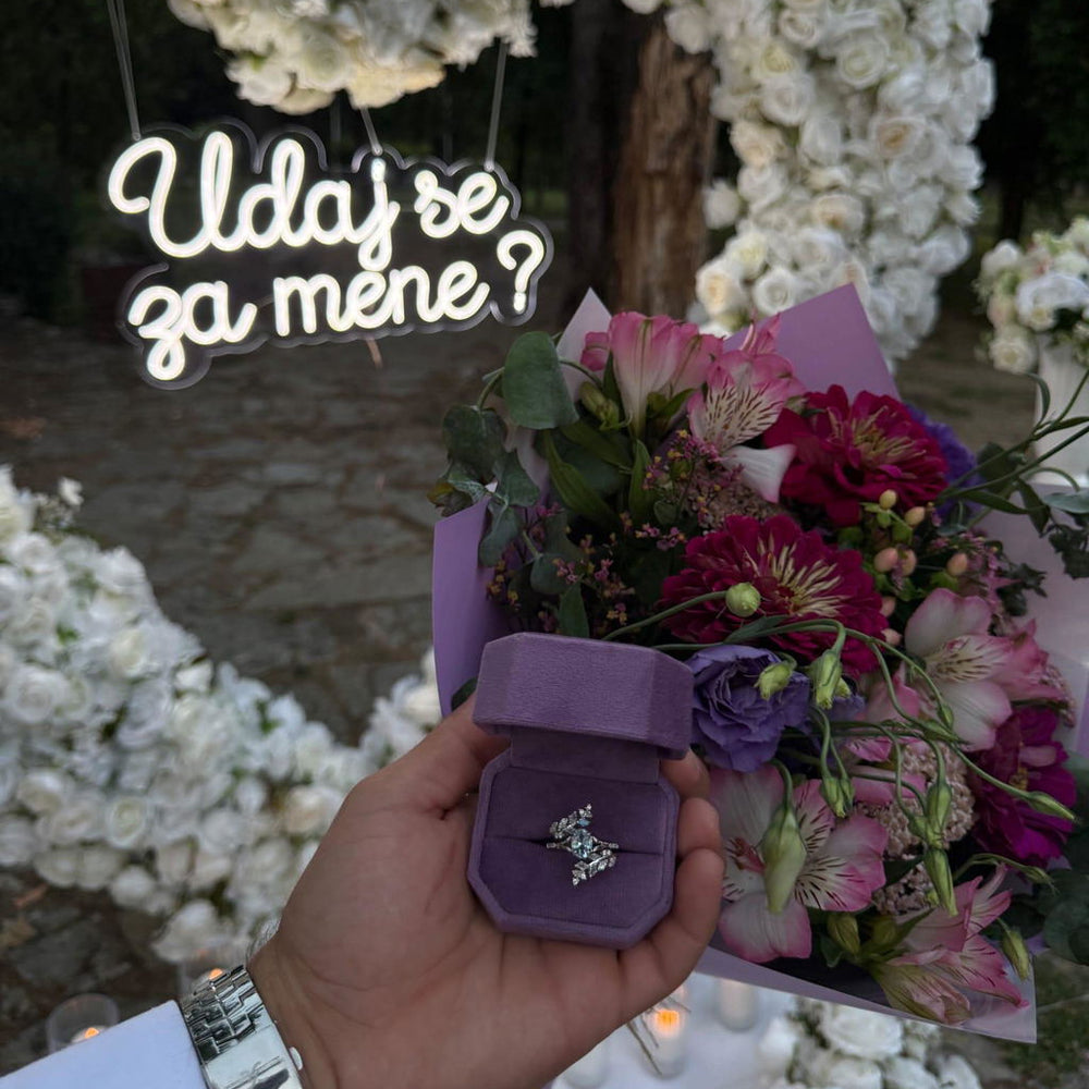 Customer proposal photo holding a lavender velvet ring box with an Aquamarise engagement ring, beside a pink bouquet and a neon marriage proposal sign