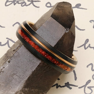 Solar Flare orange sandstone wedding band in brushed black and rose gold tungsten resting on a smoky quartz crystal