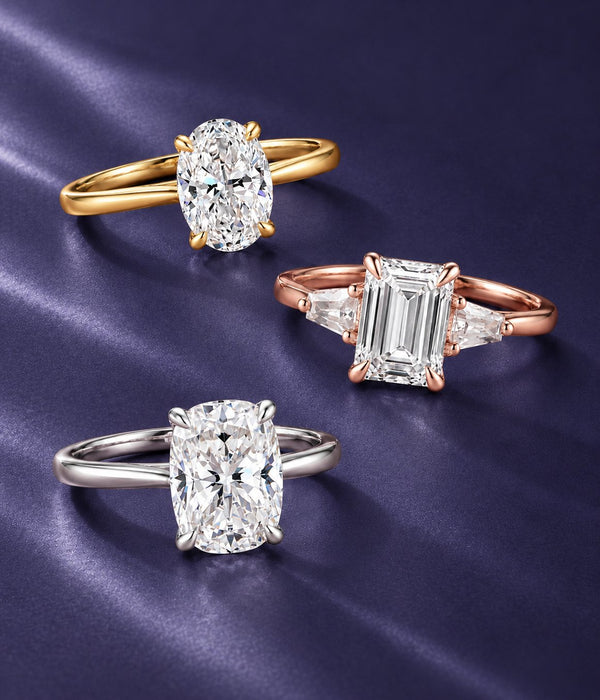 Three solitaire engagement rings on navy satin: oval in yellow gold, emerald-cut three-stone in rose gold and cushion-cut in white gold, Aquamarise banner