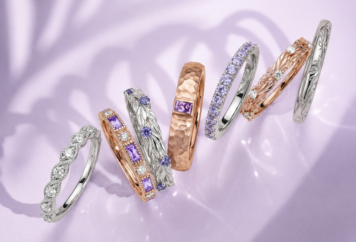 Wedding bands by Aquamarise fanned across soft lilac fabric, including rose gold, yellow gold and rhodium-finished styles set with diamonds and violet gemstones.